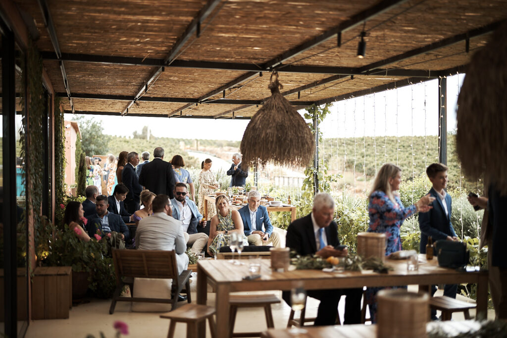 Atmosphärisches Hochzeitsfoto von Gästen auf der überdachten Terrasse des Arvad in der Algarve während des Empfangs mit Gesprächen, Drinks und Blick in die Landschaft.