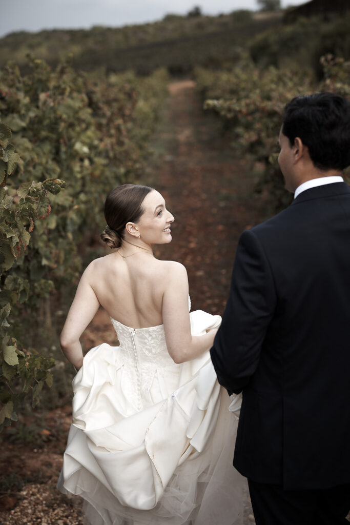 Braut blickt lächelnd über ihre Schulter während sie mit dem Bräutigam durch den Weinberg auf dem Weingut Arvad geht