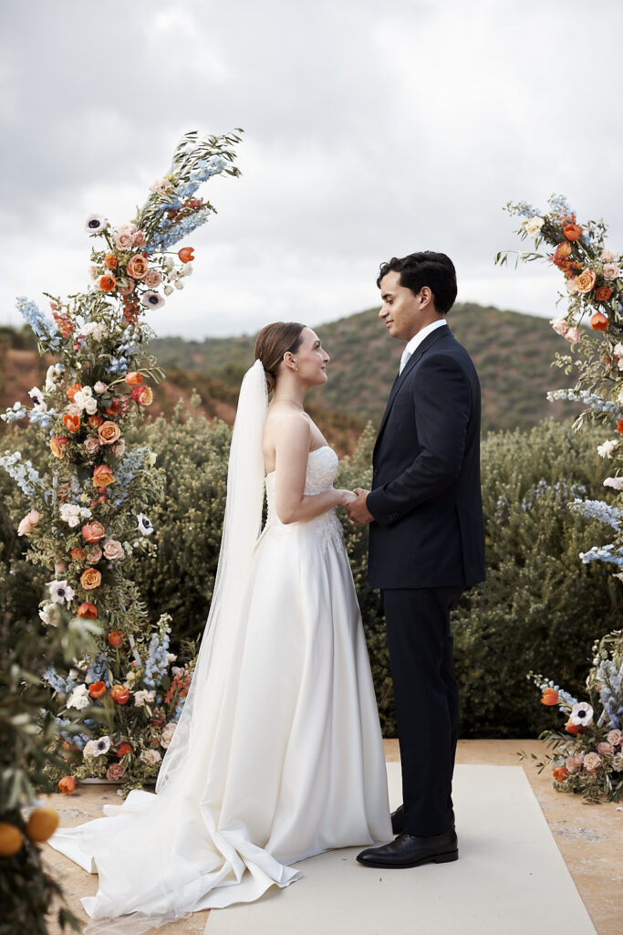Gefühlvolle Hochzeitsaufnahme des Ringtausches auf dem Weingut Arvad in der Algarve. Braut und Bräutigam stehen unter dem farbenreichen Blumenbogen, während sie sich während der freien Zeremonie die Ringe anstecken.