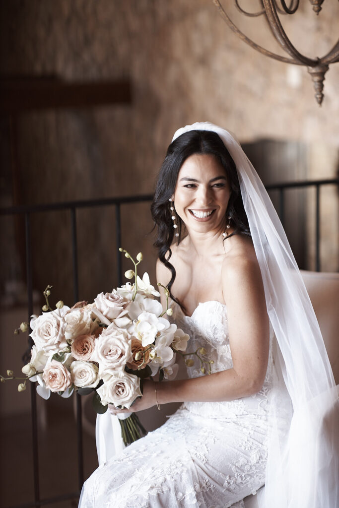 Braut mit Schleier und Brautstrauß sitzt lächelnd während der Vorbereitungen im Monte do Serrinho an der Algarve, Portugal