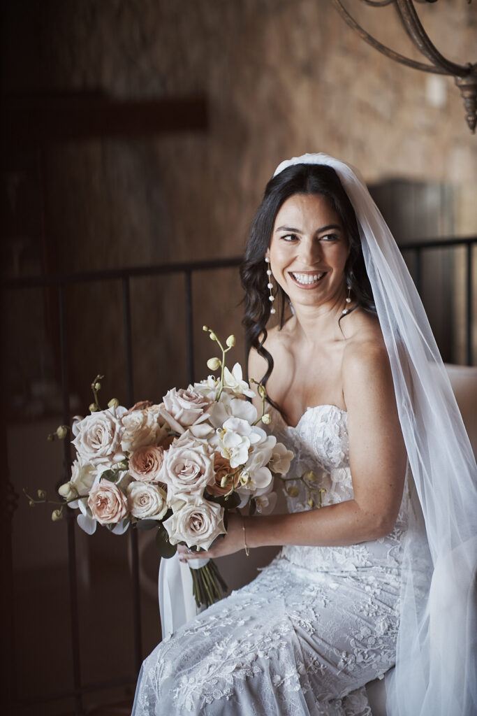 Elegantes Brautporträt während der Hochzeitsvorbereitungen im Monte do Serrinho an der Algarve, Portugal. Die Braut sitzt im Innenbereich mit langem Schleier und einem Brautstrauß aus Rosen und Orchideen.