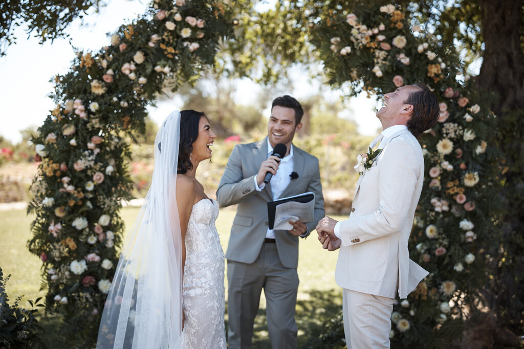Braut und Bräutigam lachen während der Trauung unter dem Blumenbogen im Monte do Serrinho an der Algarve, Portugal