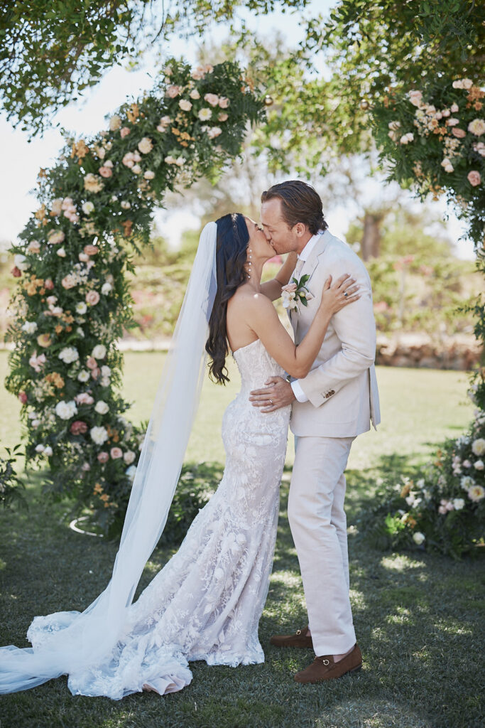 Braut und Bräutigam küssen sich nach der Trauung unter einem Blumenbogen bei ihrer Monte do Serrinho Hochzeit an der Algarve