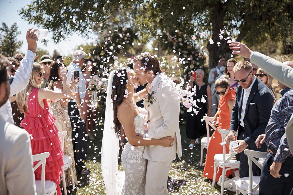Braut und Bräutigam küssen sich nach der Trauung zwischen den Gästen und fliegenden Blütenblättern im Monte do Serrinho an der Algarve