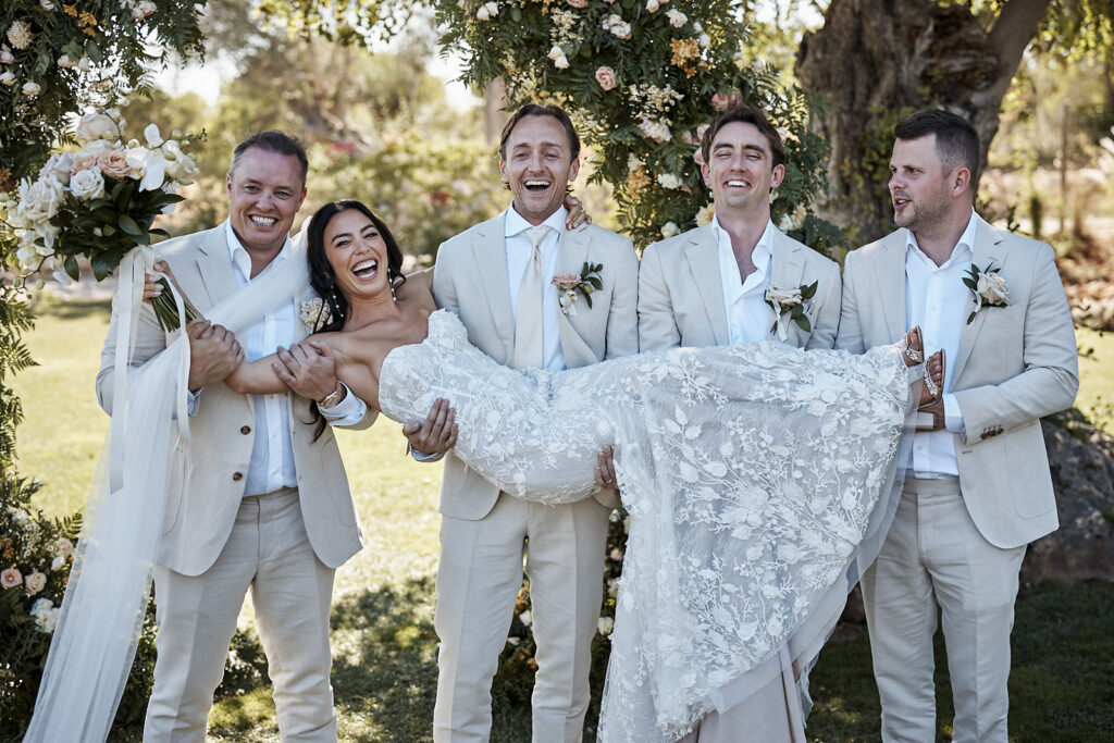 Braut wird lachend von Bräutigam und Groomsmen unter dem Blumenbogen bei der Monte do Serrinho Hochzeit an der Algarve getragen