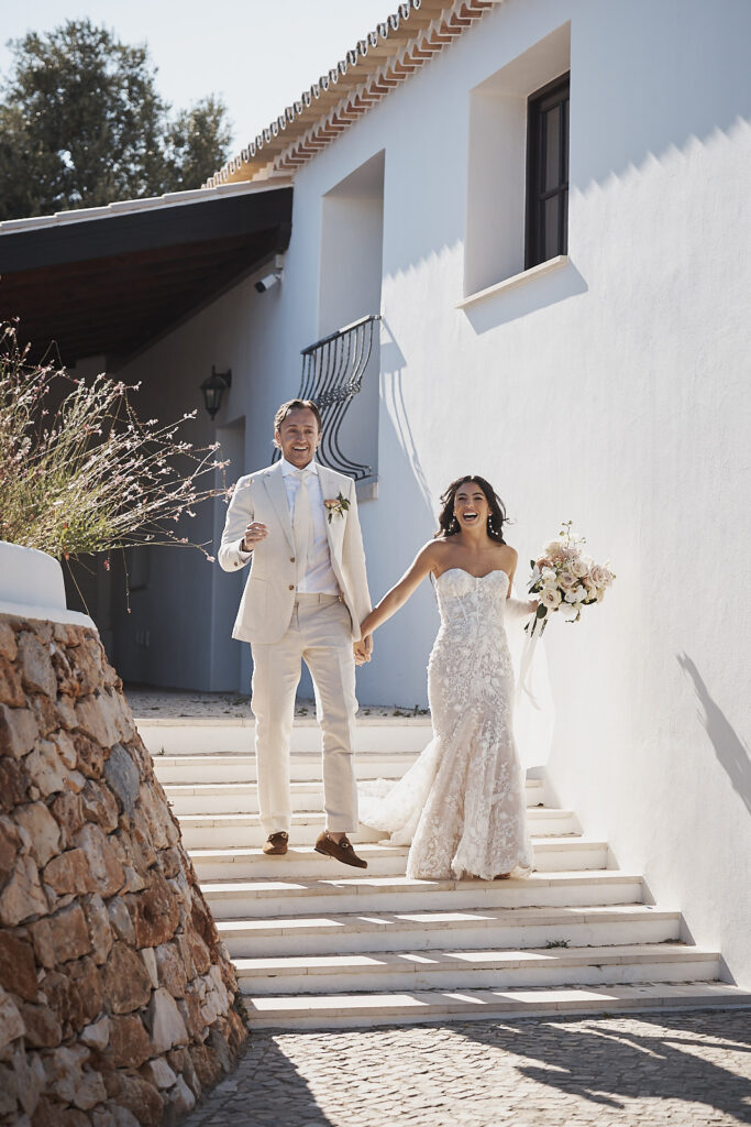 Braut und Bräutigam laufen Hand in Hand eine weiße Treppe bei ihrer Hochzeit im Monte do Serrinho an der Algarve hinunter