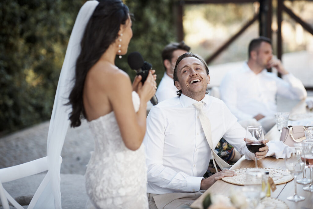 Braut hält eine Rede mit Mikrofon beim Dinner, während der Bräutigam im Monte do Serrinho an der Algarve lächelnd zuhört
